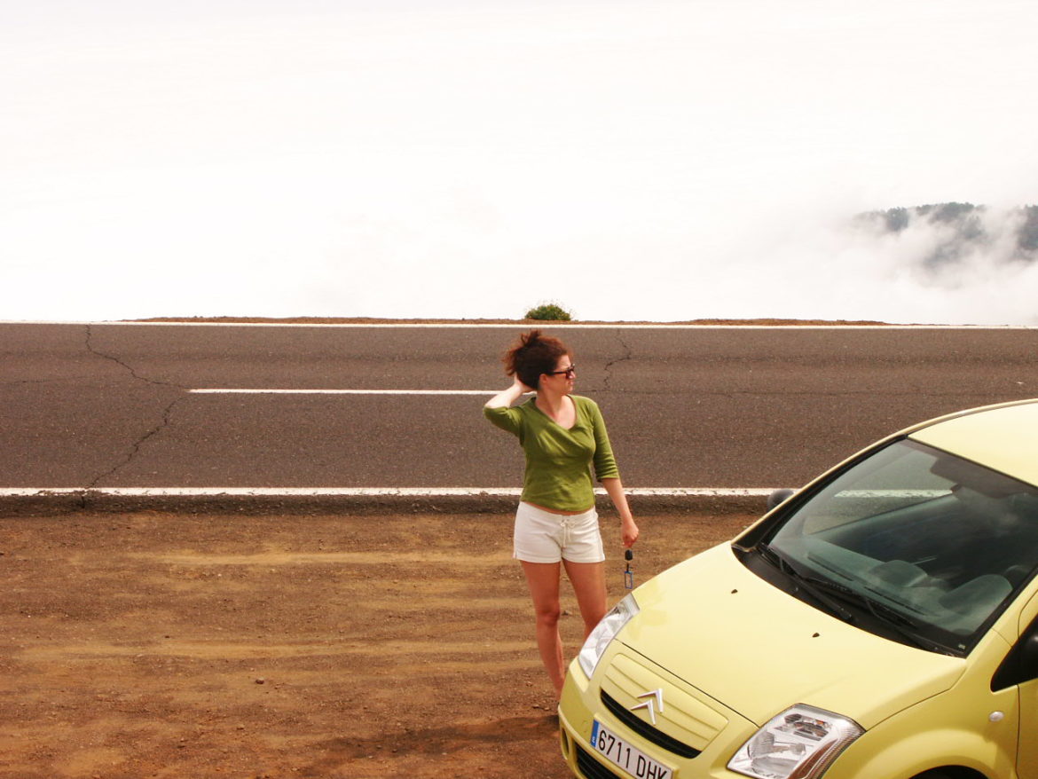 Schlanke Frau mit Autoschlüssel hinter einem gelben Citroen kratzt sich am Hinterkopf. Dunkler Asphalt dahinter und Nebel zieht auf.