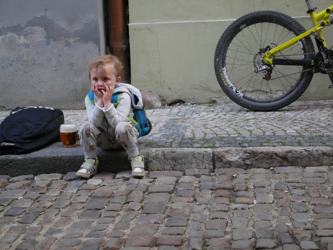 Verzweifeltes Kind sitzt am Gehsteigrand, neben ihm steht ein volles Bierglas und ein Fahrrad. Prag