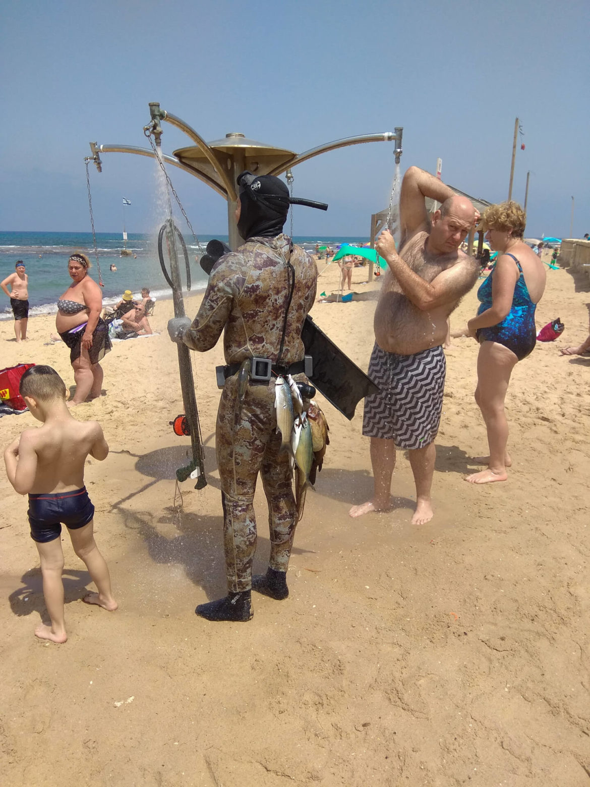Israel Reisebericht. Sporttaucher im Neoprenanzug mit Harpune und gefangenen Fischen, welche am Hintern hängen duscht sich am Strand
