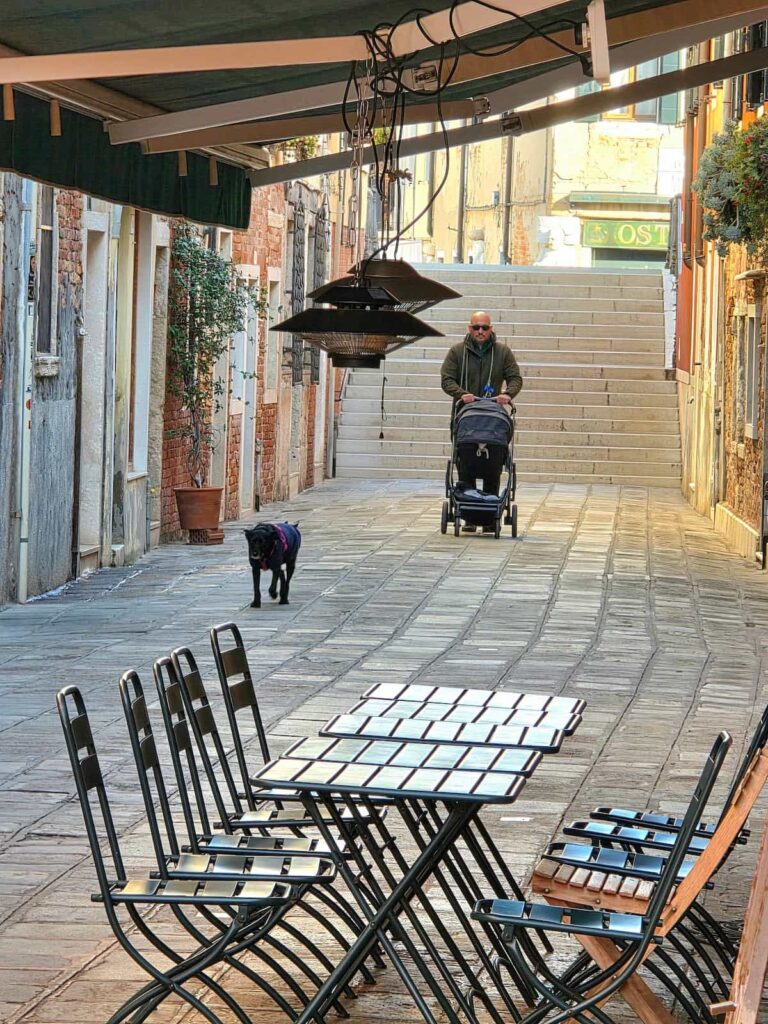 Vater mit Kinderwagen und schwarzem Hund in einer Gasse in Venedig
